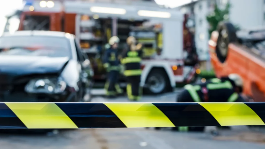 Firefighters working near a fire truck, with a yellow and black caution tape in the foreground, indicating an emergency scene